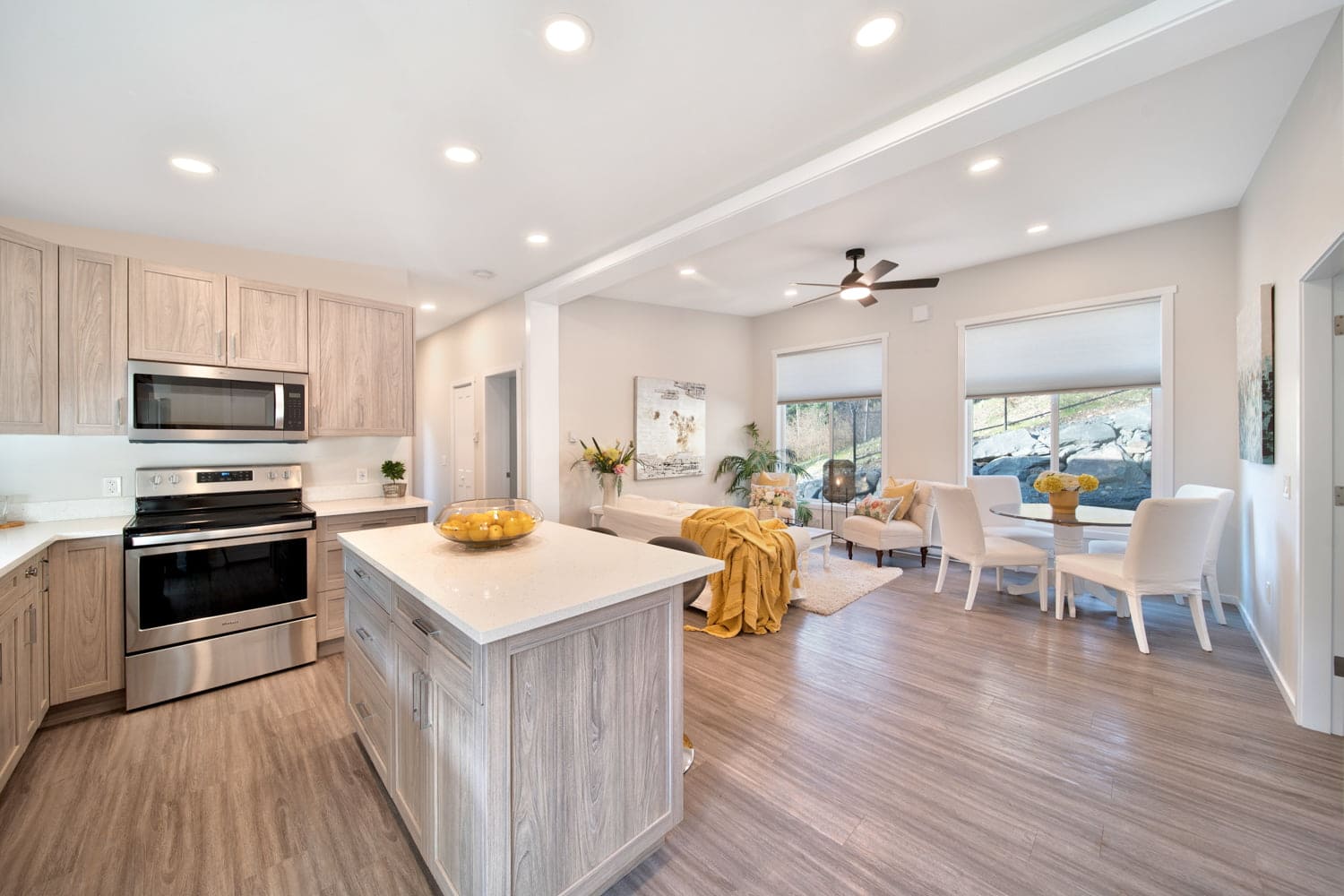 Bright open kitchen with center island, stainless steel appliances, and view into a furnished living and dining area.